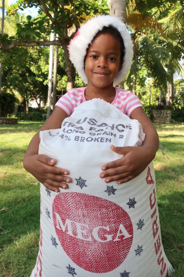 A girl in a santa hat holding a big bag of Rice for Christmas Fund.