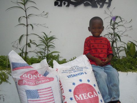 A young boy sitting in front of a wall with a Bag of Rice and Beans for Your Sponsor Child.