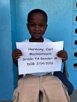 A young boy holding up a sign that reads harmoly carl mackencour grade p gender m.