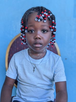 A young girl with dreadlocks sitting in front of a blue wall.