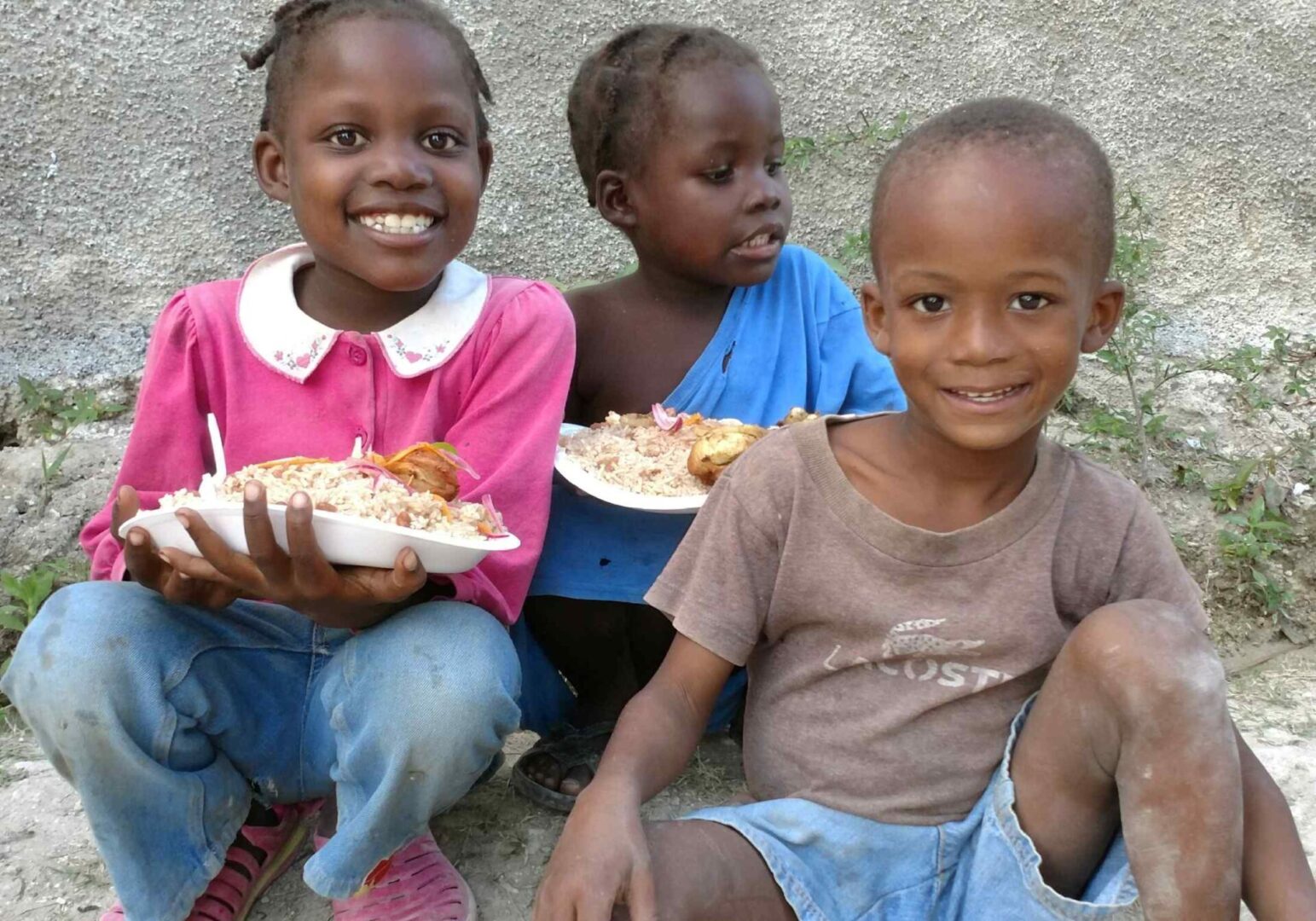 Three children sitting on the ground with plates of food.